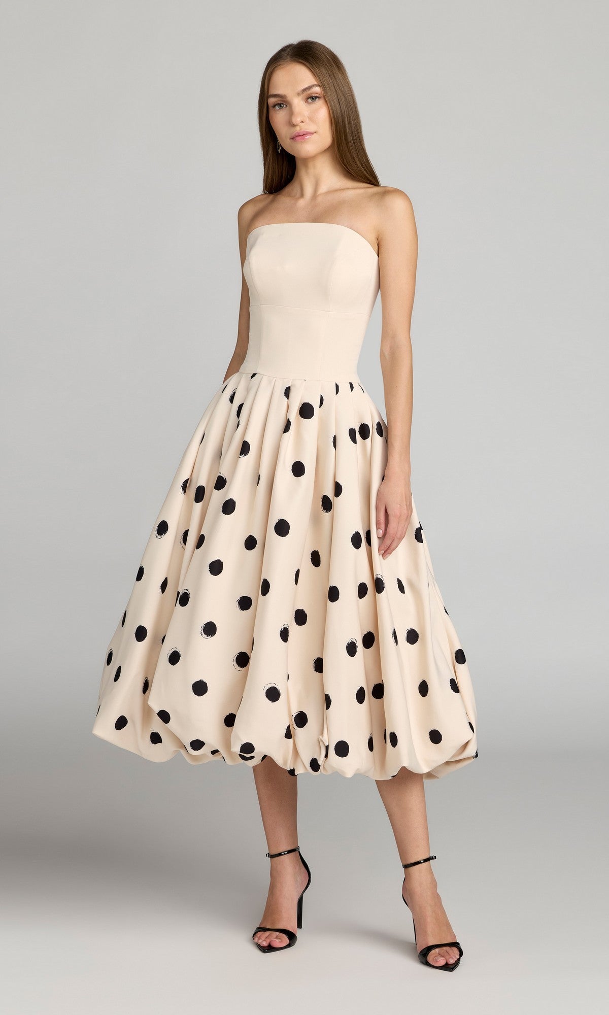 A woman wears the Mac Duggal 21234 Semi-Formal Polka Dot Midi Dress in Ecru Multi—featuring a strapless bodice, full A-line skirt, and cream hues—elegantly paired with black high-heeled sandals.